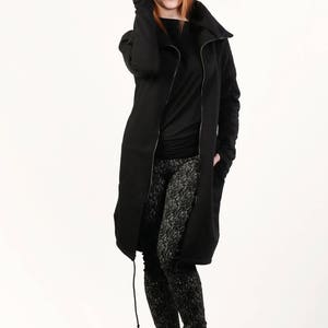May include: A black long-sleeved jacket with a zipper closure and a high collar. The jacket has a drawstring at the bottom and is worn over a black long-sleeved shirt and black and white patterned leggings. The person is wearing black ankle boots and a black fedora.