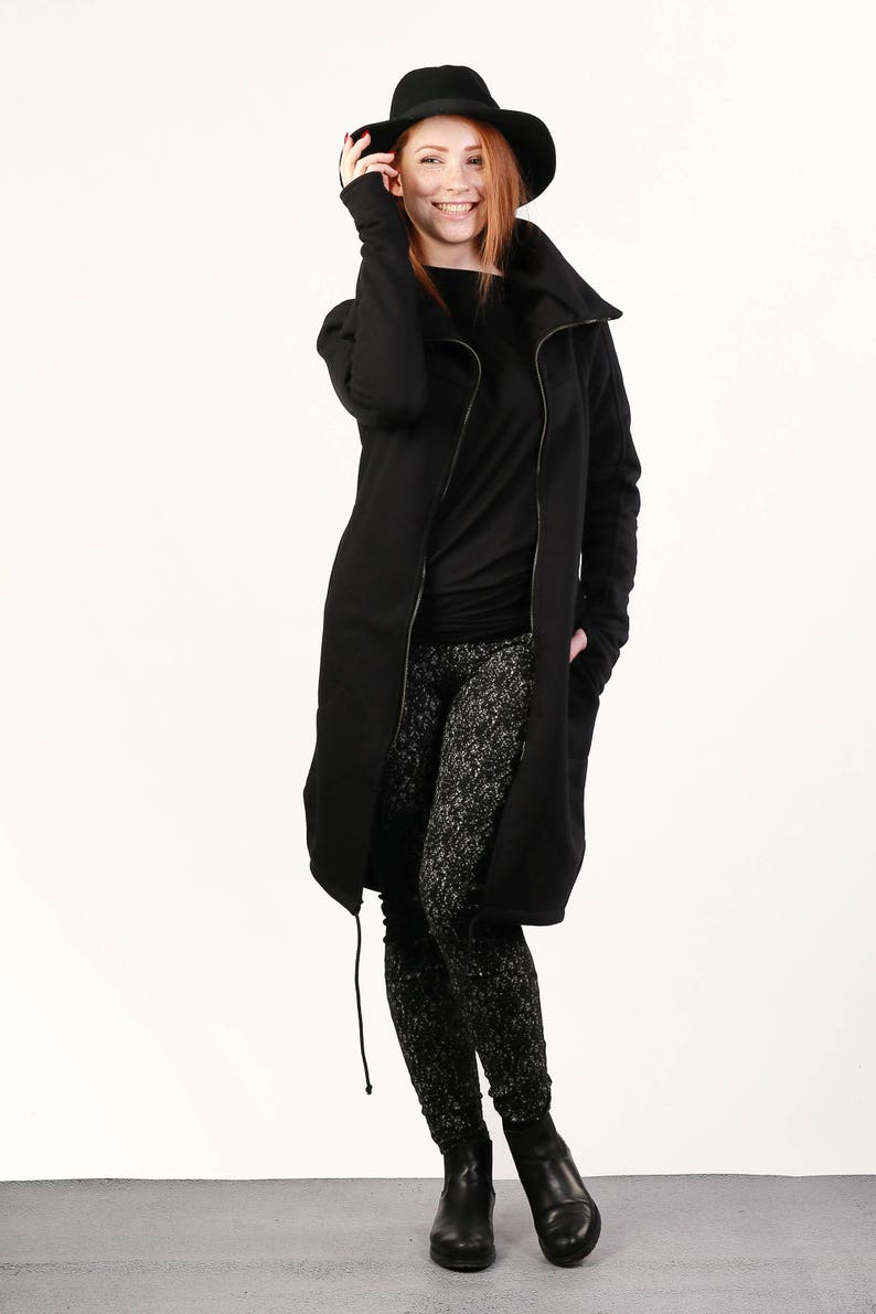 May include: A black long-sleeved jacket with a zipper closure and a high collar. The jacket has a drawstring at the bottom and is worn over a black long-sleeved shirt and black and white patterned leggings. The person is wearing black ankle boots and a black fedora.