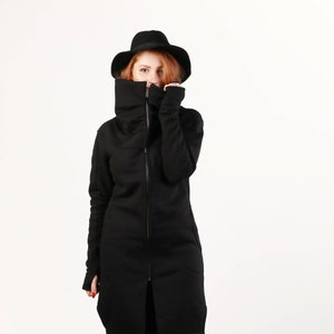 Könnte beinhalten: Eine schwarze Jacke mit langen Ärmeln, einem hohen Kragen und einem Reißverschluss. Die Jacke hat eine lockere Passform und eine lange Länge. Das Model trägt einen schwarzen Hut.