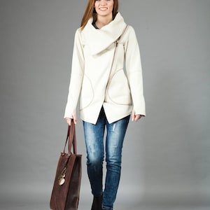 May include: A woman wearing a white asymmetrical zippered jacket with a brown leather tote bag. She is wearing blue jeans and black ankle boots.