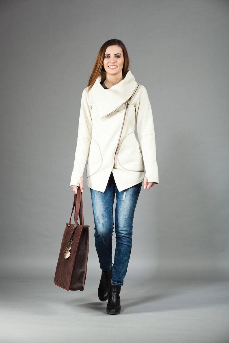 May include: A woman wearing a white asymmetrical zippered jacket with a brown leather tote bag. She is wearing blue jeans and black ankle boots.