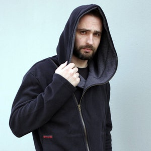 May include: A black zip-up hoodie featuring a large, deep hood with a contrasting grey interior lining. The garment includes a full-length front zipper, long sleeves, and a small rectangular red label detail on the lower front pocket area.