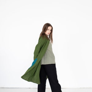May include: A woman wearing a long, green linen coat with a blue patch on the sleeve. She is also wearing a light green shirt and black pants.