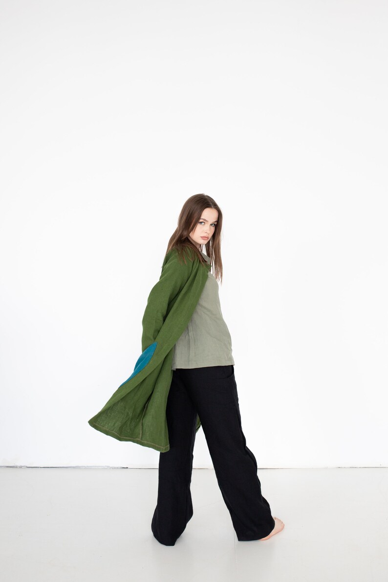 May include: A woman wearing a long, green linen coat with a blue patch on the sleeve. She is also wearing a light green shirt and black pants.