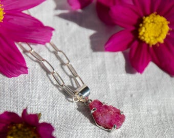 May include: A silver chain necklace with a pendant featuring a raw pink ruby gemstone. The pendant is set in silver and has a delicate design. The necklace is photographed on a white fabric background with pink flowers.