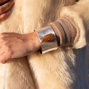 May include: A wide, silver cuff bracelet with a large, irregularly shaped, amber-coloured stone. The bracelet is worn on a wrist, and the background is a cream-coloured, fuzzy coat.