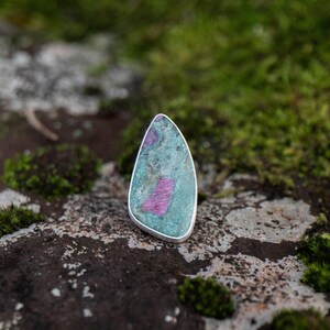 May include: A teardrop-shaped pendant featuring a polished surface with a mottled green and grey base colour, accented by a distinct, rectangular-shaped magenta inclusion set within a smooth, thin silver-toned metal border frame.