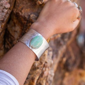 May include: A wide, silver cuff bracelet with a large, oval, light green stone. The bracelet is worn on a wrist, and the background is a blurred view of a rock formation. The cuff has a textured surface.