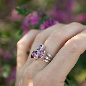 May include: A silver ring with three oval and square-cut, deep pink gemstones. The ring has a textured band with two horizontal lines. The gemstones are set in a bezel setting. The ring is displayed on a hand.