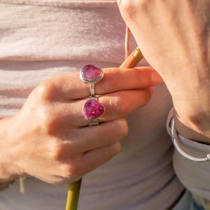 May include: Two silver rings featuring teardrop-shaped, textured magenta-pink stones. The rings are worn on adjacent fingers, showcasing a metallic band design with a polished finish and vibrant, deep pink gemstone settings that catch the light.