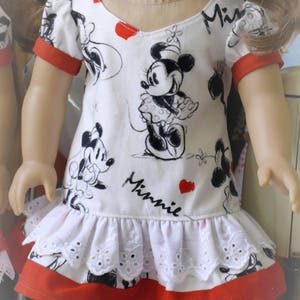 May include: A doll wearing a white dress with red trim and a black and white Minnie Mouse print. The dress has a white lace ruffle at the hem. The doll is wearing white sandals.