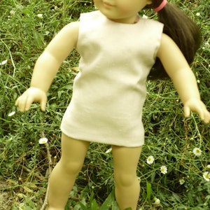 Pode incluir: Uma boneca de menina pequena com cabelo castanho comprido vestindo um vestido branco sem mangas. A boneca está em pé em um campo de grama verde e flores brancas.