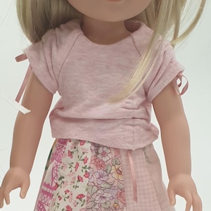 May include: A doll wearing a light pink t-shirt with short sleeves and a skirt. The skirt is a patchwork design with floral and geometric patterns. The doll has blonde hair.