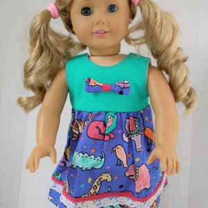 May include: A doll wearing a teal sleeveless dress with a purple and pink bow. The dress has a blue skirt with a white lace trim and a colourful animal print.