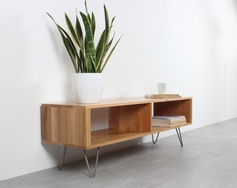 May include: A rectangular light wood media console featuring two open storage compartments and four slender silver metal hairpin legs. A white ceramic pot containing a tall green snake plant sits on top, alongside a small white cup and books.