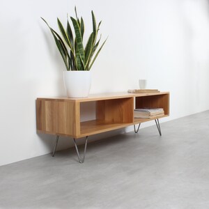 May include: A rectangular light wood media console featuring two open storage compartments and four thin, silver-colored metal hairpin legs. A white ceramic pot containing a tall green snake plant sits on the left side of the top surface.