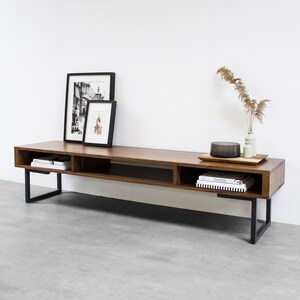 May include: A long, dark brown wooden TV stand with a black metal frame. The stand has three shelves and is decorated with two black framed pictures and a vase with dried flowers.