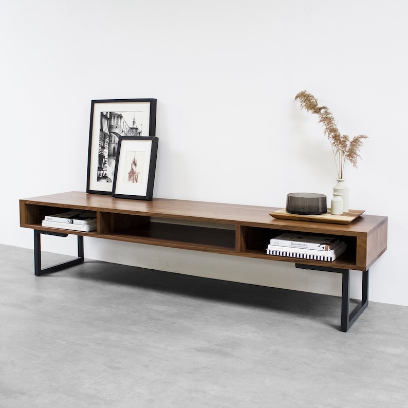 May include: A long, dark brown wooden TV stand with a black metal frame. The stand has three shelves and is decorated with two black framed pictures and a vase with dried flowers.