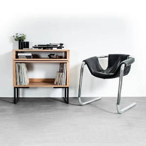 May include: A wooden record player stand with two shelves, black metal legs, and a turntable on the top shelf. The stand is next to a black leather chair with a chrome frame.