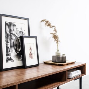 May include: A wooden entertainment center with black metal legs. The center has two shelves and a drawer. There are two framed black and white drawings and a vase with dried flowers on top of the center.