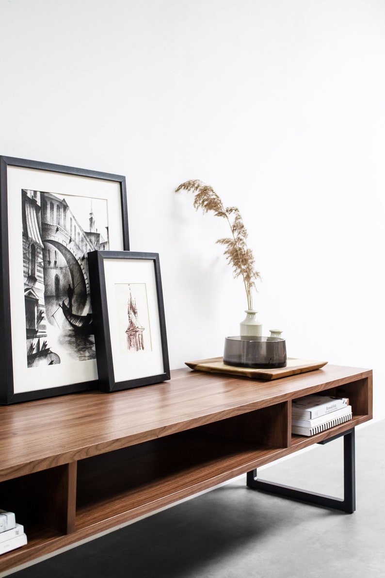 May include: A wooden entertainment center with black metal legs. The center has two shelves and a drawer. There are two framed black and white drawings and a vase with dried flowers on top of the center.