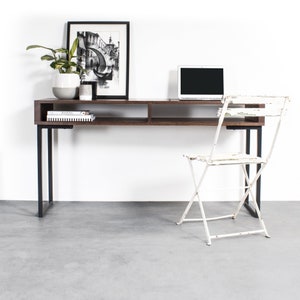 May include: A dark brown wooden desk with a black metal frame and a white metal folding chair. The desk has a shelf for storage and a laptop computer on the surface. There is a framed black and white drawing of a building on the wall behind the desk.