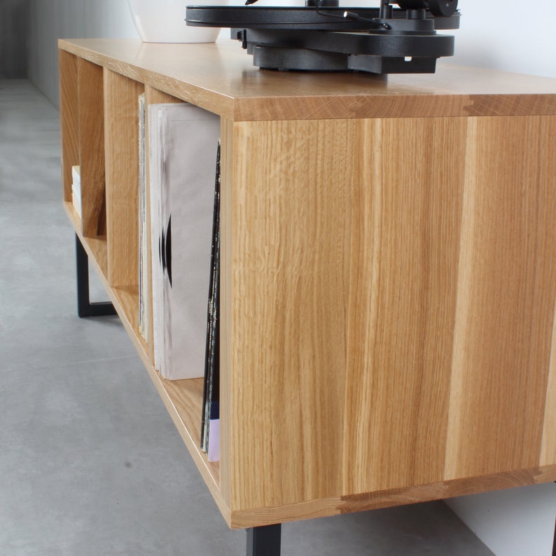 Puede incluir: Un soporte para tocadiscos de madera clara con patas de metal negro. El soporte tiene cuatro estantes para guardar discos de vinilo.