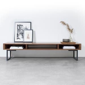 May include: A long, dark brown wooden media console with black metal legs. The console has two open shelves and is decorated with framed black and white photos and a vase with dried grasses.
