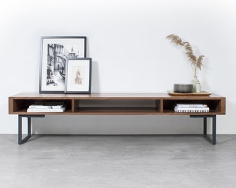 May include: A long, dark brown wooden media console with black metal legs. The console has two open shelves and is decorated with framed black and white photos and a vase with dried grasses.