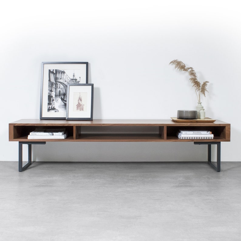 May include: A long, dark brown wooden media console with black metal legs. The console has two open shelves and is decorated with framed black and white photos and a vase with dried grasses.