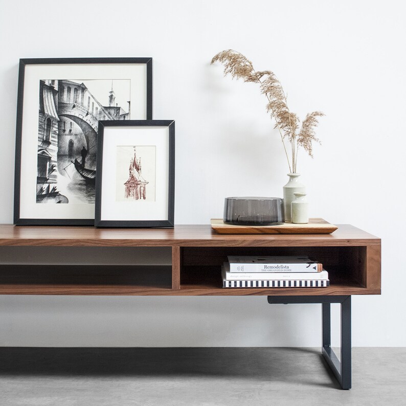 May include: A dark brown wooden media console with black metal legs. The console has two open shelves and a closed shelf. There are two framed black and white drawings on the top shelf, a clear glass bowl, and a stack of books on the bottom shelf. A vase with dried reeds is on the right side of the console.