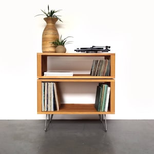 May include: A two-tiered wooden shelf unit with hairpin legs, featuring a turntable on the top shelf and vinyl records and books on the lower shelf. The shelf is made of natural wood and has a light brown finish.