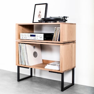 May include: A light oak record player stand with black metal legs. The stand has two open shelves, one holding a silver stereo receiver and vinyl records. A framed picture and a black turntable sit on top.