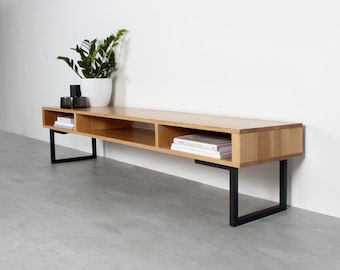May include: A long, rectangular media console crafted from light-toned wood, featuring three open storage compartments. The unit is supported by two black metal U-shaped legs. The surface holds a white potted plant and a dark glass container.