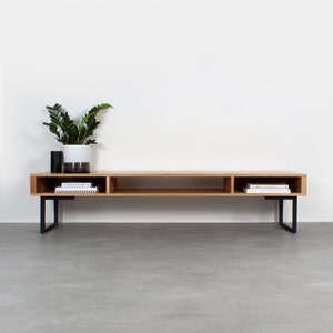 May include: A long, rectangular light wood media console featuring three open storage compartments and two black metal U-shaped legs. A white and dark gray ceramic planter with a green plant sits on the left side of the top surface.