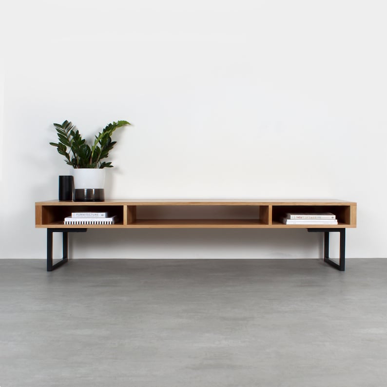 May include: A long, rectangular light wood media console featuring three open storage compartments and two black metal U-shaped legs. A white and dark gray ceramic planter with a green plant sits on the left side of the top surface.