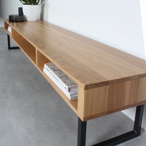 May include: A long, rectangular media console made of light-toned wood with a smooth grain finish. The unit features open shelving compartments and is supported by a black metal frame with rectangular legs. Books are stored inside the shelves.