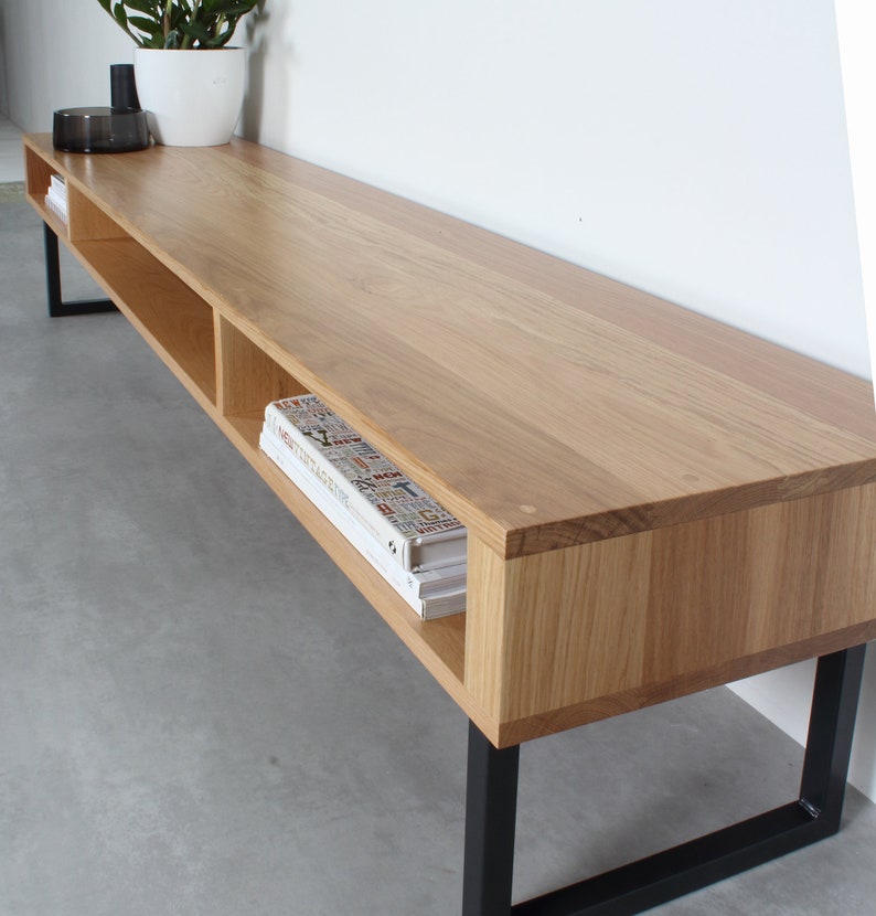 May include: A long, rectangular media console made of light-toned wood with a smooth grain finish. The unit features open shelving compartments and is supported by a black metal frame with rectangular legs. Books are stored inside the shelves.