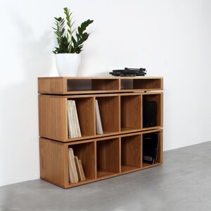 May include: A three-tiered wooden record shelf with a turntable on top. The shelf has 12 compartments for storing records. The shelf is made of light brown wood and has a modern design.