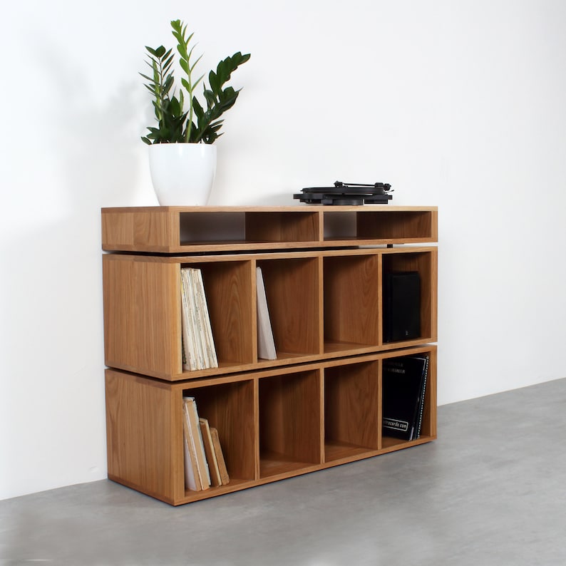 May include: A three-tiered wooden record shelf with a turntable on top. The shelf has 12 compartments for storing records. The shelf is made of light brown wood and has a modern design.