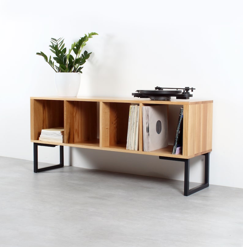 Puede incluir: Un soporte de madera para tocadiscos con cuatro estantes abiertos y patas de metal negro. El soporte est&aacute; lleno de discos de vinilo y tiene una tornamesa en la parte superior.
