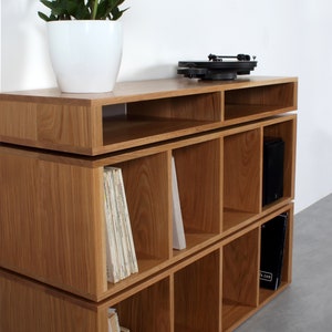 May include: A three-tiered wooden record shelf with a turntable on the top shelf. The shelf has six compartments on each level and is made of light brown wood.