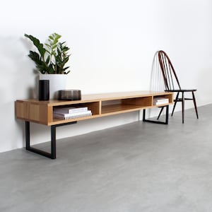 May include: A long, rectangular light wood media console featuring three open storage compartments and a smooth top surface. The unit is supported by two black metal U-shaped legs. A dark wooden spindle-back chair sits to the right of the console.