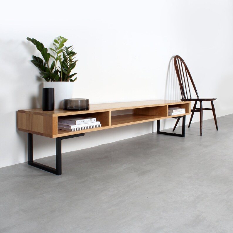 May include: A long, rectangular light wood media console featuring three open storage compartments and a smooth top surface. The unit is supported by two black metal U-shaped legs. A dark wooden spindle-back chair sits to the right of the console.