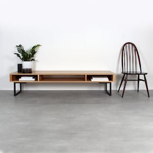 May include: A low-profile rectangular media console made of light-toned wood with three open storage compartments and black metal legs. A dark brown wooden spindle-back chair sits to the right, and a potted green plant rests on the left side.