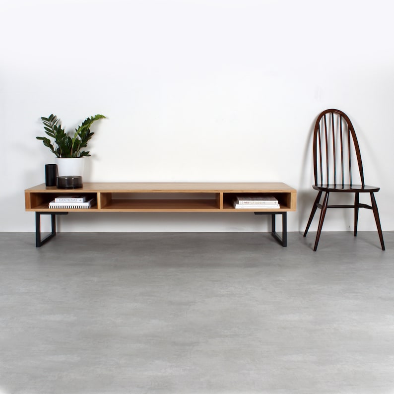 May include: A low-profile rectangular media console made of light-toned wood with three open storage compartments and black metal legs. A dark brown wooden spindle-back chair sits to the right, and a potted green plant rests on the left side.