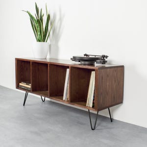 May include: A dark brown wooden media console with four open storage compartments supported by black metal hairpin legs. A black record player sits on top, and a green snake plant in a white pot is placed on the left side of the unit.