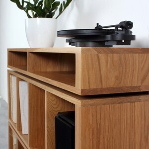 May include: A wooden record player stand with shelves for vinyl records and a speaker. The stand is made of light brown wood and has a modern design.