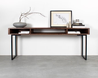 Könnte beinhalten: Ein langer, dunkelbrauner Holz-Konsolentisch mit schwarzen Metallbeinen. Der Tisch hat zwei offene Regalbretter und ein schwarz gerahmtes Bild darüber.