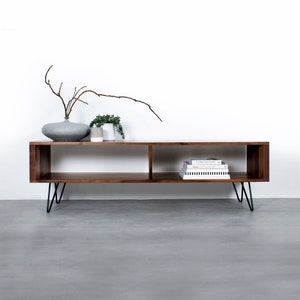 May include: A modern, rectangular wooden media console with two open compartments. The console is a warm brown color and has black hairpin legs. A gray vase with branches, small plants, and books are displayed on top.